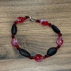 May include: A beaded bracelet featuring alternating red and black beads. The bracelet has oval-shaped black beads and round, translucent red beads. Small red and black beads connect the larger beads, with a silver clasp.