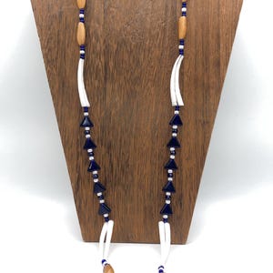 May include: A necklace with a variety of beads. The necklace features wooden, blue, and white beads, with a silver clasp. The necklace is displayed on a wooden stand against a white background.