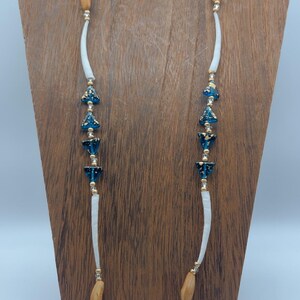 May include: A long necklace with wooden beads, gold accents, and blue floral-shaped beads. The necklace has a white, curved section on each side. The necklace is displayed on a wooden stand.
