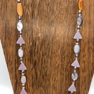 May include: A long necklace featuring a mix of wooden, oval-shaped beads and iridescent, oval-shaped beads. The necklace also includes triangular lavender beads and small silver-colored spacers. The necklace is displayed on a wooden stand.