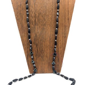 May include: A black beaded necklace with oval and round beads, interspersed with silver-colored spacers. The necklace features a silver clasp and is displayed on a wooden jewelry stand. The beads have a matte finish.