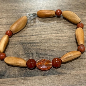 May include: A handmade bracelet featuring alternating light brown wooden beads and small, round, reddish-brown beads. The bracelet also includes a central, oval-shaped, orange and brown patterned bead, and a silver-colored clasp.