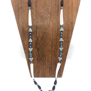 May include: A long necklace with a pendant, displayed on a wooden stand. The necklace features black oval beads, blue and gray triangular beads, and white rectangular accents. The pendant is a rectangular shape with a pearlescent finish.