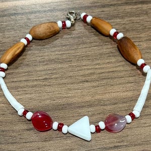 May include: A beaded bracelet featuring wooden, white, and red beads, along with red and translucent stones and a white triangular bead. The bracelet is strung on a white cord and has a silver clasp.
