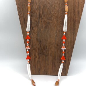 May include: A handcrafted necklace featuring wooden beads, orange accents, and white elements. The necklace has a silver clasp and is displayed on a wooden stand. The design incorporates geometric shapes and natural materials.