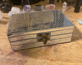 May include: A small, silver mirrored box with a brass latch. The box has a grid pattern on the top and a smooth, mirrored surface on the sides.