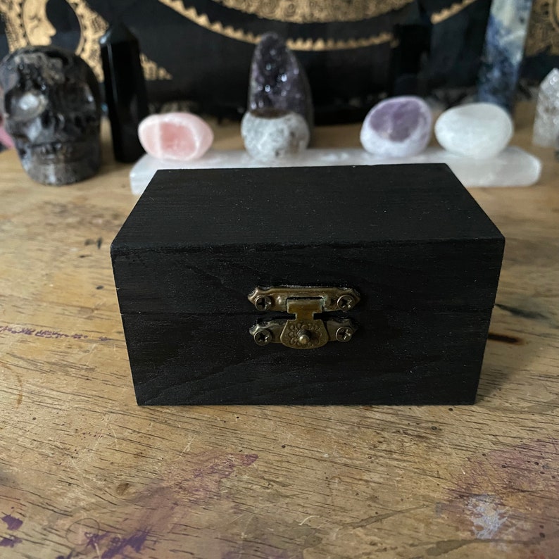 May include: A small black wooden box with a brass latch. The box is closed and sitting on a wooden surface.