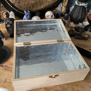 May include: A light-colored wooden box with a mirrored interior and lid, open to reveal the reflective surface. The box has brass hinges and a clasp. The background includes various decorative objects.