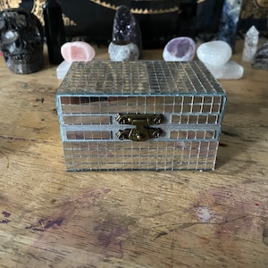 May include: A small, silver mirrored box with a brass latch. The box has a tiled design and is closed.