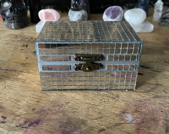 May include: A small, silver mirrored box with a brass latch. The box has a tiled design and is closed.