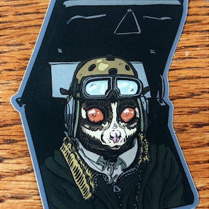 May include: A sticker featuring a cartoon illustration of a lemur wearing a pilot's helmet and goggles, sitting in the cockpit of an aircraft. The lemur has large, orange eyes and a pink nose. The background is a dark blue and black color.