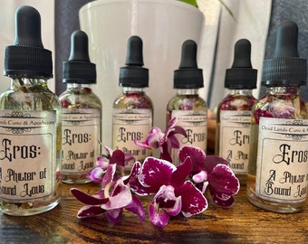 May include: Six small glass bottles with dropper tops filled with a clear liquid and dried flowers. The bottles have labels that read "Eros: A Phiuter of Sound Love". The bottles are arranged in a row on a wooden surface with a purple orchid flower in the foreground.