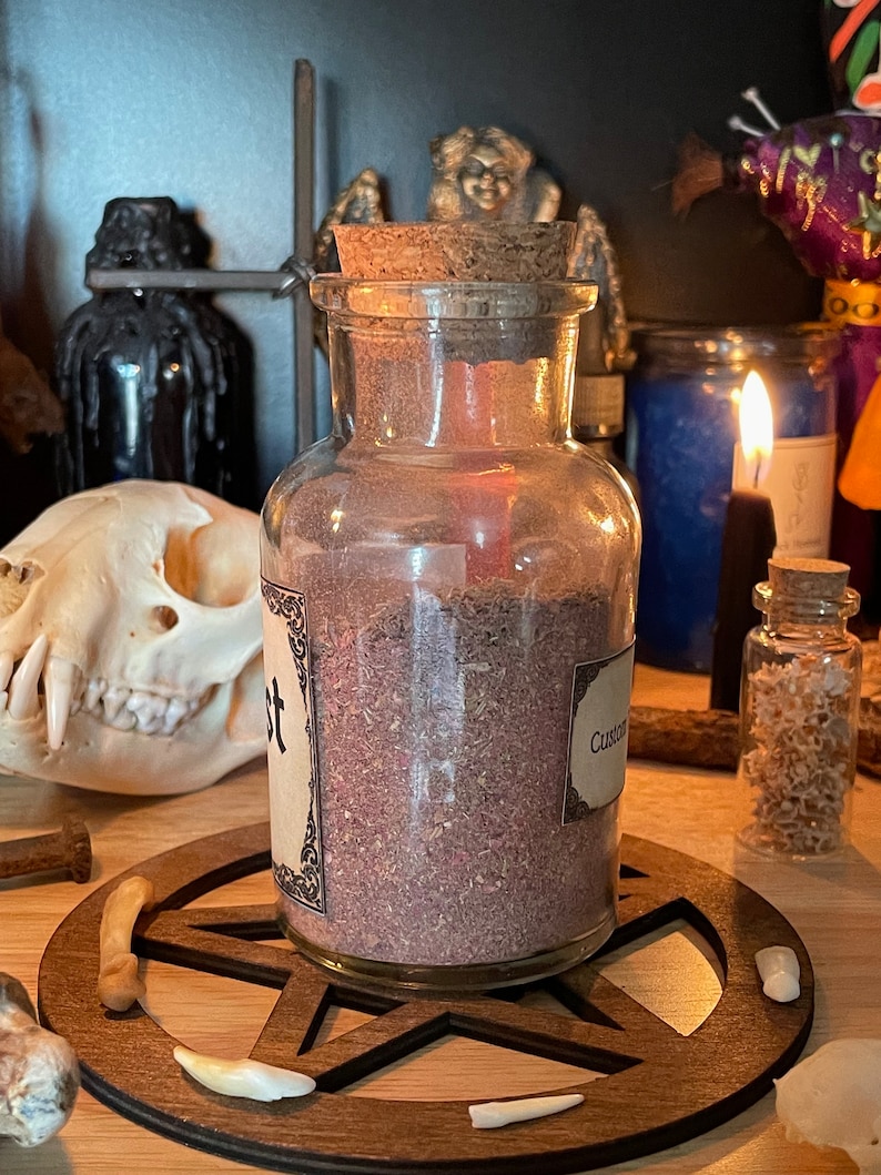 May include: A glass jar with a cork lid filled with a purple powder. The jar is labeled "Custom Blend". The jar is sitting on a wooden pentagram.
