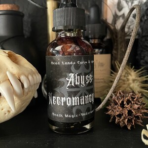 May include: A small glass bottle with a black dropper top and a black label. The label reads "Dead Lands Curio & Apothecary Abyss Necromancy Death Magic - Underworld".