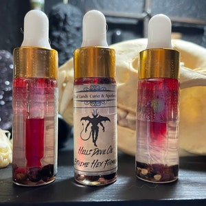 May include: Three small glass bottles with gold lids and droppers filled with a red liquid and small black and brown objects. The bottles have white labels with black text that reads "Old Lands Curio & Apothecary" and "Hells Devil Oil Extreme Hex Formula".