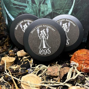 May include: Three black round containers with white labels that read "Dead Lands Apothecary" and feature a witch with a pointed hat and a staff. The middle container has the year "1693" printed on the label.