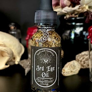 May include: A small glass bottle with a black dropper top filled with a clear liquid and herbs. The label reads "Dead Lands Apothecary 3rd Eye Oil Black Cumin Seed Decadility".