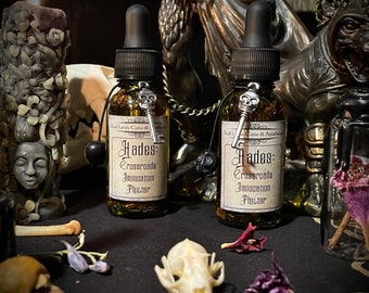 May include: Two glass bottles with dark liquid and black droppers. Each bottle has a label that reads "Hades: Crossroads Invocation Phuter." A silver key charm with a skull hangs from each bottle. The bottles are part of a collection of apothecary items.