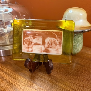 May include: A rectangular yellow glass plaque featuring a centered white rectangular insert with a brown-toned ultrasound image of a fetus. The glass has two small metal loops at the top and is displayed on a dark brown wooden stand.