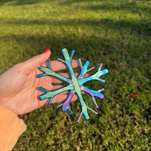 May include: A multi-colored snowflake ornament with blue, teal, and purple hues. The ornament has a glittery finish and is held in a hand against a backdrop of green grass and a wooden fence.