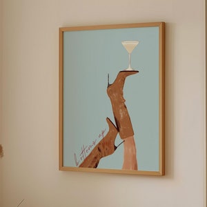 May include: A framed print with a blue background featuring a pair of brown boots with a martini glass on top. The text "bottoms up" is written in pink on the print.
