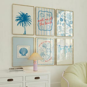 May include: Six framed beach-themed art prints with gold frames. The prints feature blue illustrations of a palm tree, seashells, and the text "meet me at the beach." A white cabinet with a lamp and a book are in the foreground.