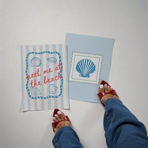 May include: Two cards with beach-themed designs. One card has blue and white stripes with seashells and the text "meet me at the beach." The other card is light blue with a seashell stamp. Red sandals and blue jeans are also visible.