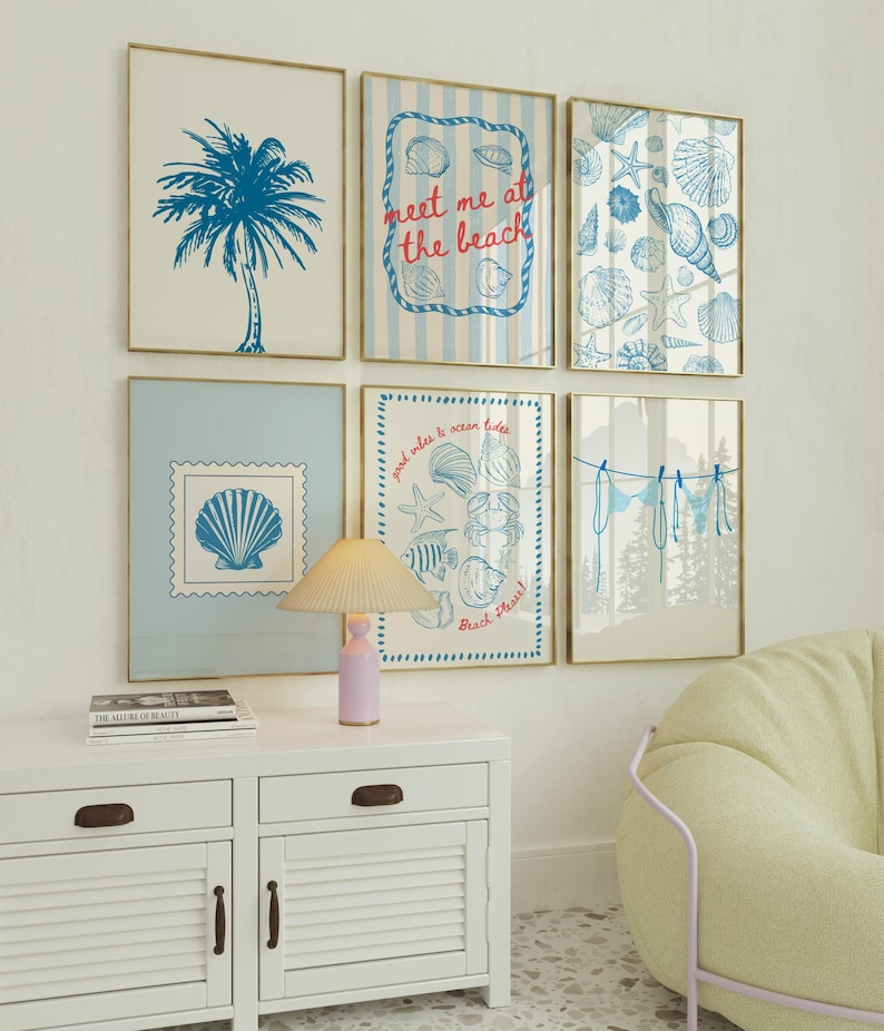 May include: Six framed beach-themed art prints with gold frames. The prints feature blue illustrations of a palm tree, seashells, and the text "meet me at the beach." A white cabinet with a lamp and a book are in the foreground.