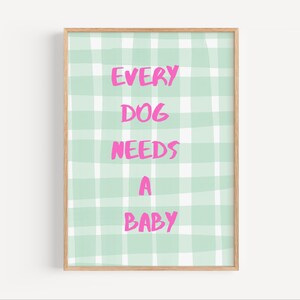 Op de afbeelding: Een ingelijste print met de tekst "EVERY DOG NEEDS A BABY" in felroze letters. De achtergrond is een lichtgroen en wit ruitpatroon. De lijst is lichtbruin.