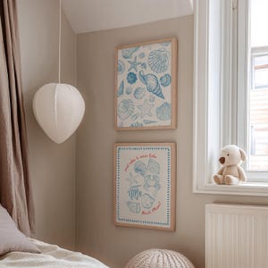 May include: Two framed prints with seashell illustrations in blue and white hang on a beige wall. The top print features various shells and starfish. The bottom print has text that reads "good vibes & ocean tides" and "Beach Please!"