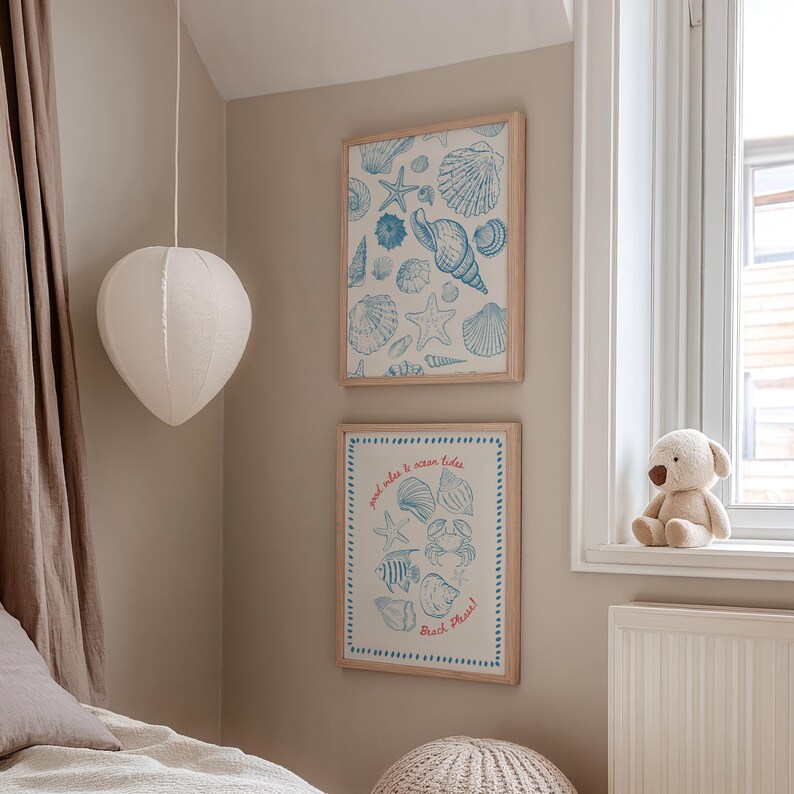 May include: Two framed prints with seashell illustrations in blue and white hang on a beige wall. The top print features various shells and starfish. The bottom print has text that reads "good vibes & ocean tides" and "Beach Please!"