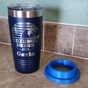 May include: A navy blue stainless steel tumbler with a silver lid and a white engraved design featuring palm trees and the text "Cozumel Mexico 2023 Gavin". The tumbler is sitting on a brown tiled surface next to a blue silicone lid.