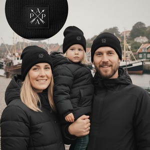 Könnte beinhalten: Eine dreiköpfige Familie trägt passende schwarze Mützen und Jacken. Die Mützen haben ein kleines quadratisches Patch mit den Buchstaben "P A P A" und Pfeildesigns. Die Familie steht in der Nähe eines Hafens mit Booten.