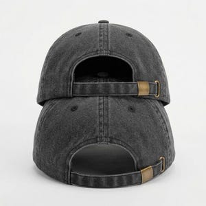 May include: Two dark gray baseball caps stacked on top of each other. The caps have a distressed, worn look. Each cap has a curved brim and an adjustable strap with a brass-colored buckle on the back.