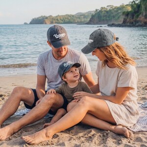 May include: A family of three wearing matching gray baseball caps with the words "Vintage" and "Original" printed on them. They are sitting on a sandy beach with the ocean in the background. The caps are a faded gray color.