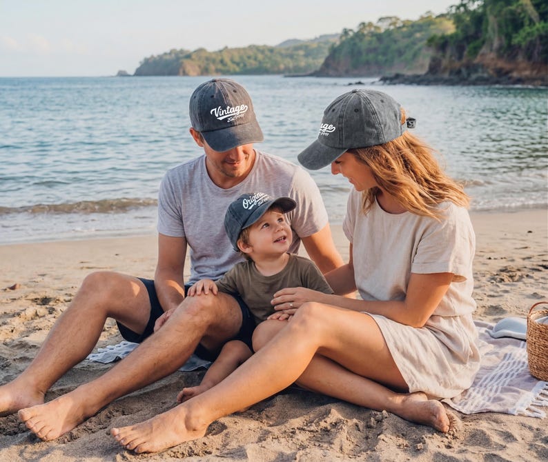 May include: A family of three wearing matching gray baseball caps with the words "Vintage" and "Original" printed on them. They are sitting on a sandy beach with the ocean in the background. The caps are a faded gray color.