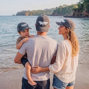 May include: A family at the beach wearing matching gray vintage baseball caps. The caps have white text that reads "Original" and "Vintage Est. 1993". The family is standing near the water's edge.