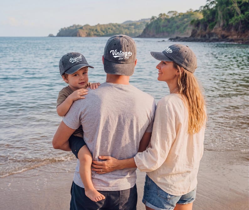 May include: A family at the beach wearing matching gray vintage baseball caps. The caps have white text that reads "Original" and "Vintage Est. 1993". The family is standing near the water's edge.