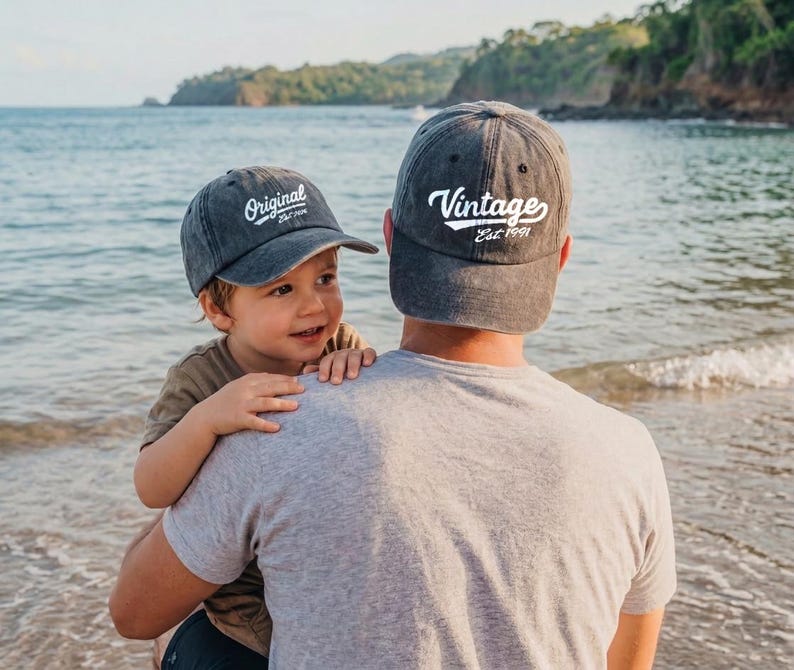May include: Two gray baseball caps with white text, one reading "Original Est. 1991" and the other "Vintage Est. 1991." A child is wearing the "Original" cap, and an adult is wearing the "Vintage" cap. The caps are worn at the beach.