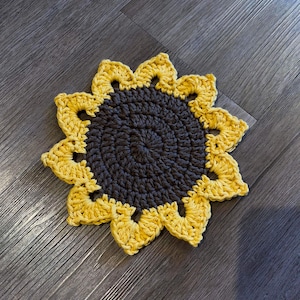 Può includere: Un sottopentola a forma di girasole lavorato a uncinetto, con un centro marrone e petali gialli.