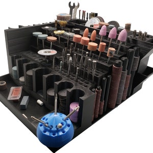 May include: A black, multi-tiered organizer filled with various rotary tool bits, sanding drums, and accessories. The organizer includes a blue tool holder and a wrench. The tools are in various shapes and sizes, including cylindrical, conical, and disc-shaped.