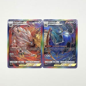 Puede incluir: Dos cartas coleccionables holográficas que presentan a Solgaleo GX y Lunala GX. La carta de Solgaleo muestra una criatura blanca similar a un león con detalles rojos y dorados, mientras que la de Lunala muestra una criatura celestial azul y morada con un borde arcoíris.