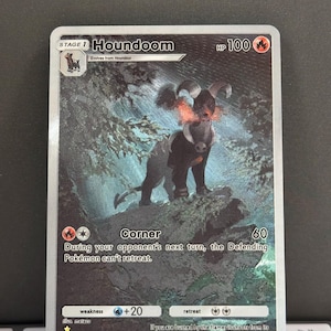 Puede incluir: Una carta de Pokémon que presenta a Houndoom, una criatura oscura con forma de perro y acentos de fuego. La carta muestra el nombre "Houndoom", 100 PS y detalles de ataque. El fondo muestra una escena oscura y boscosa.
