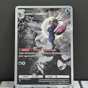 Puede incluir: Una carta coleccionable de Pokémon Greninja. La carta presenta una ilustración en blanco y negro de Greninja con un diseño rosa y amarillo. La carta tiene el texto "Water Shuriken" y "Mist Slash".