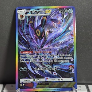 以下が含まれることがあります： ホログラフィックデザインのUmbreon EXポケモンカード。カードには、黄色のアクセントが付いた濃い紫と黒のUmbreonのキャラクターが描かれています。カードには虹色のボーダーがあり、「Dark Chase」や「Darkness Fang」などのテキストが含まれています。