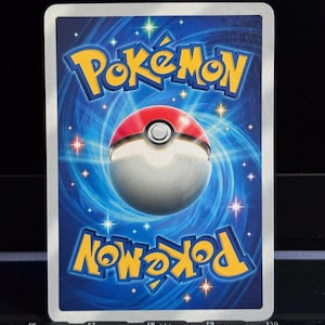 Puede incluir: El reverso de una carta coleccionable de Pok&eacute;mon. La tarjeta presenta un fondo azul con la palabra "Pok&eacute;mon" en amarillo en la parte superior e inferior. Una Pok&eacute; Ball roja y blanca est&aacute; en el centro.