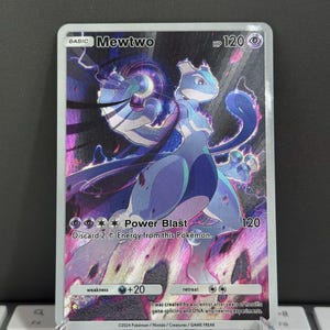 Puede incluir: Carta coleccionable Pokémon Mewtwo holográfica. La carta presenta un personaje Mewtwo morado y blanco con el texto "Power Blast" y "HP 120". La carta se muestra en un soporte transparente.