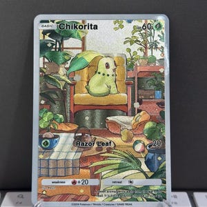 Puede incluir: Carta de juego Pokémon Chikorita con una criatura verde y amarilla en una habitación con plantas. La carta incluye "Razor Leaf" y "HP 60". La ilustración detallada muestra varias plantas y un ambiente acogedor.