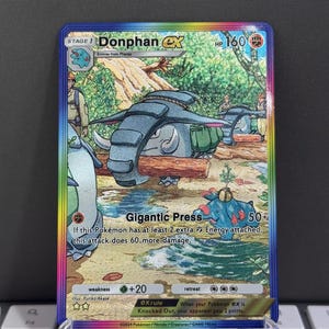 May include: A Donphan ex Pokémon trading card with a colorful border. The card features a blue and gray elephant-like creature in a forest setting. Text includes "Gigantic Press" and "HP 160".