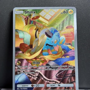 Puede incluir: Una carta de Pokémon Shinx con una ilustración azul y amarilla. La carta presenta el texto "Shinx" y "HP 60". La carta tiene un efecto holográfico y está sobre un fondo colorido.